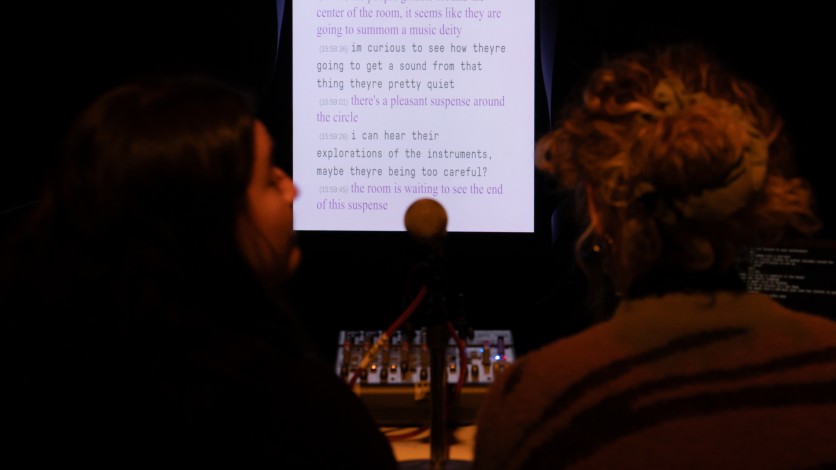Two people sitting in front of the microphone under a dimmed light. In the background, the screen shows the text that has been typed by the narrators.