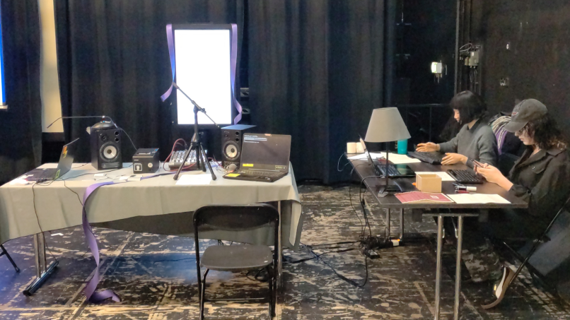 A phot of my group’s setup: a table on the left faces a vertical screen, on the tables are mics, speakers, laptop and a mixer. Another table on the right welcomes the narrators, they have keyboards, laptops, papersheets and a lamp.